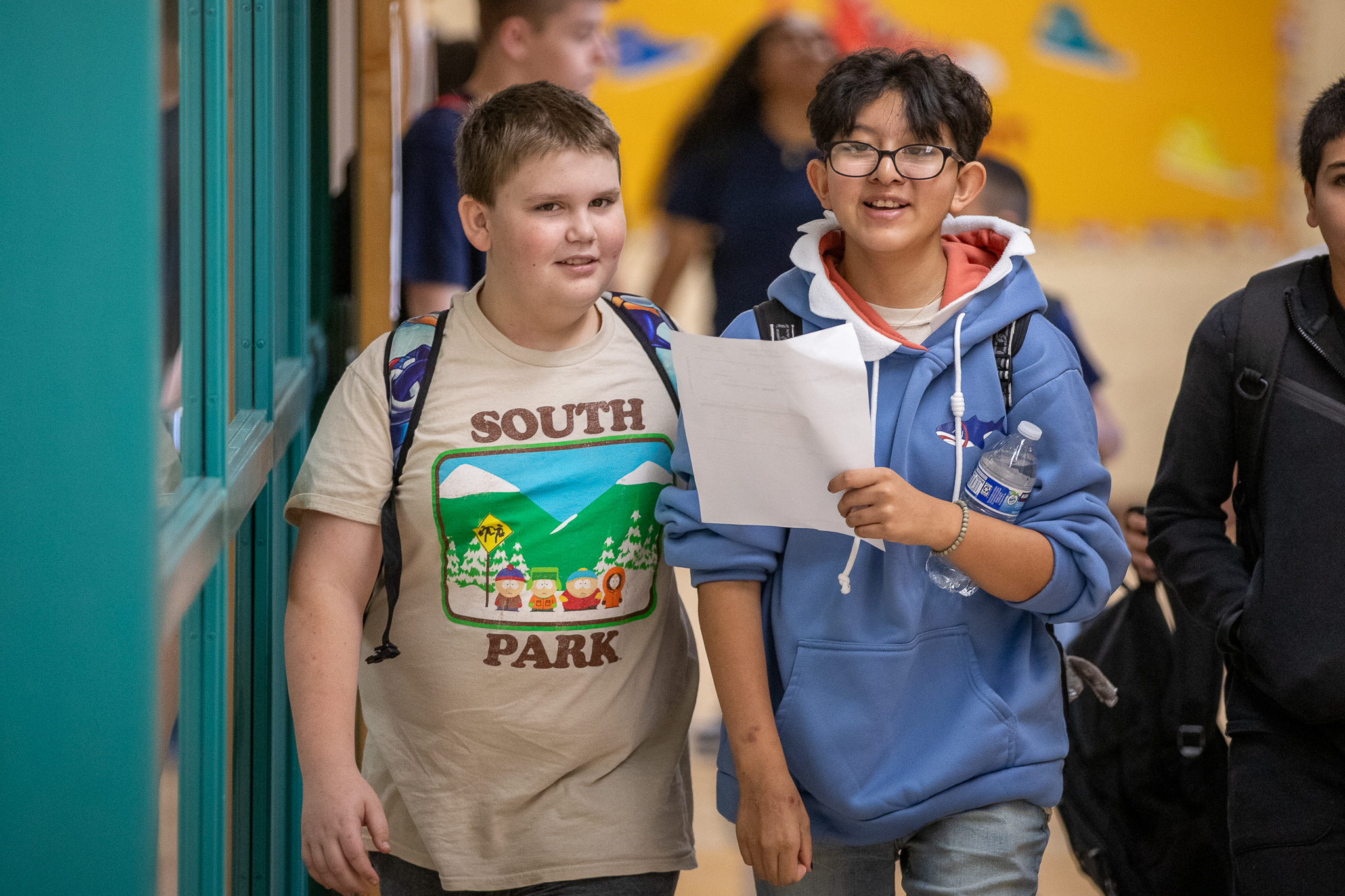 Two boys smile as they look at a piece of paper while walking in the hallway